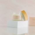 a 4 oz frosted glass jar with a very light creamy color for the product. Resting against the right side of the jar is a gold looking cap. The jar and cap are sitting on top of a white riser with the reflection seen at the bottom of the photo in front of the riser. Body Butter - Restore