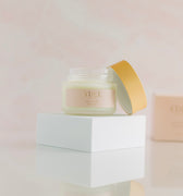 a 4 oz frosted glass jar with a very light creamy color for the product. Resting against the right side of the jar is a gold looking cap. The jar and cap are sitting on top of a white riser with the reflection seen at the bottom of the photo in front of the riser. Body Butter - Restore