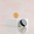 a glass jar with cream foundation sitting on a white riser with a black cap sitting at the bottom of the riser.