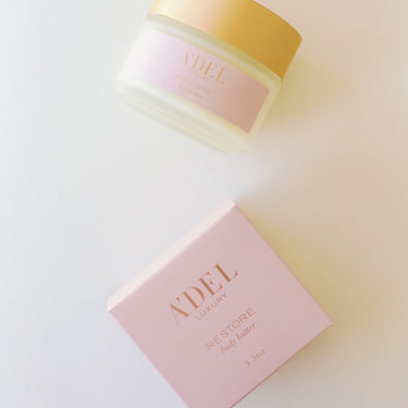 a 4 oz frosted glass jar with a very light creamy color for the product. A gold colored cap is screwed on top. Jar is laying on it's side and just below it is the light pink box that reads  Body Butter - Restore along with the A'del Logo