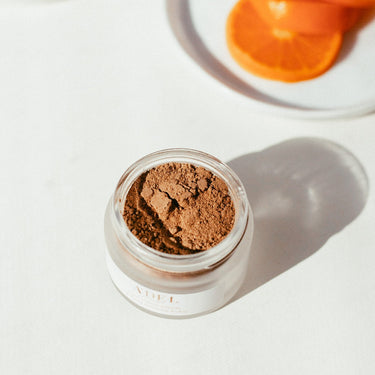 The top view of a frosted glass jar filled with a brown coffee/chocolate colored powder. Above the jar you can see a few orange slices sitting on a small plate. C&C Mask