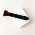 A black makeup brush with brown bristles that come to a flat, blunt shape on top. The bristles are medium to dense packed. The Brush is sitting on top of a white block with a cream background seen around it. 