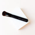 This entire brush is shown laying on the background of a white block and cream colored backdrop.A black brush with dark brown bristles. Short bristles tightly packed make up the chubby blender