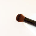 A white background with the top half of a black makeup brush with brown bristles that come to a dome shape on top. The bristles are medium to dense packed. 