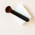 A black makeup brush with brown bristles that come to a dome shape on top. The bristles are medium to dense packed. The Brush is sitting on a white block on top of a cream background. 