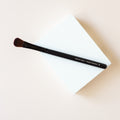 The entire brush is seen sitting on a white block with a cream background. The brush is black with dark brown bristles. The bristles seem to be in a flat line with a slight rounding on the top. The bristles are packed with a medium density and appears to look slightly fluffy.