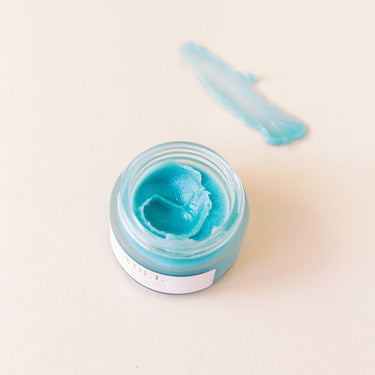 an over top view looking into the face balm jar where you see a vibrant blue balm with a finger size dent in the middle. Just beside the jar a swatch of the cream is spread showing how transparent the cream becomes when spread. Blue Lagoon Face Balm